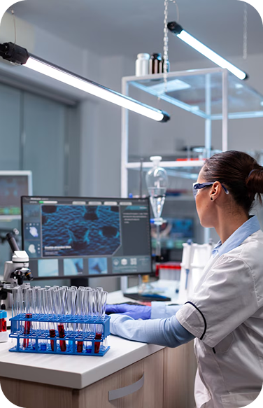 Female lab worker at a desk analyzing a computer screen with test samples in tubes next to her