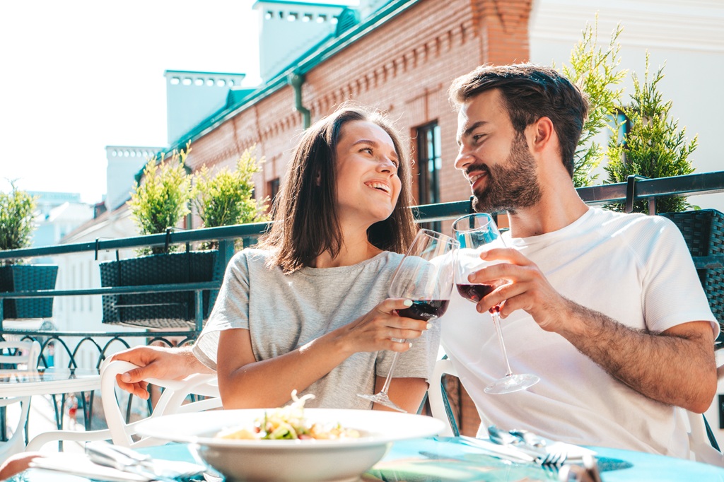 Man and woman sitting at a restaurant table outside, clinking their glasses of red wine and looking at each other