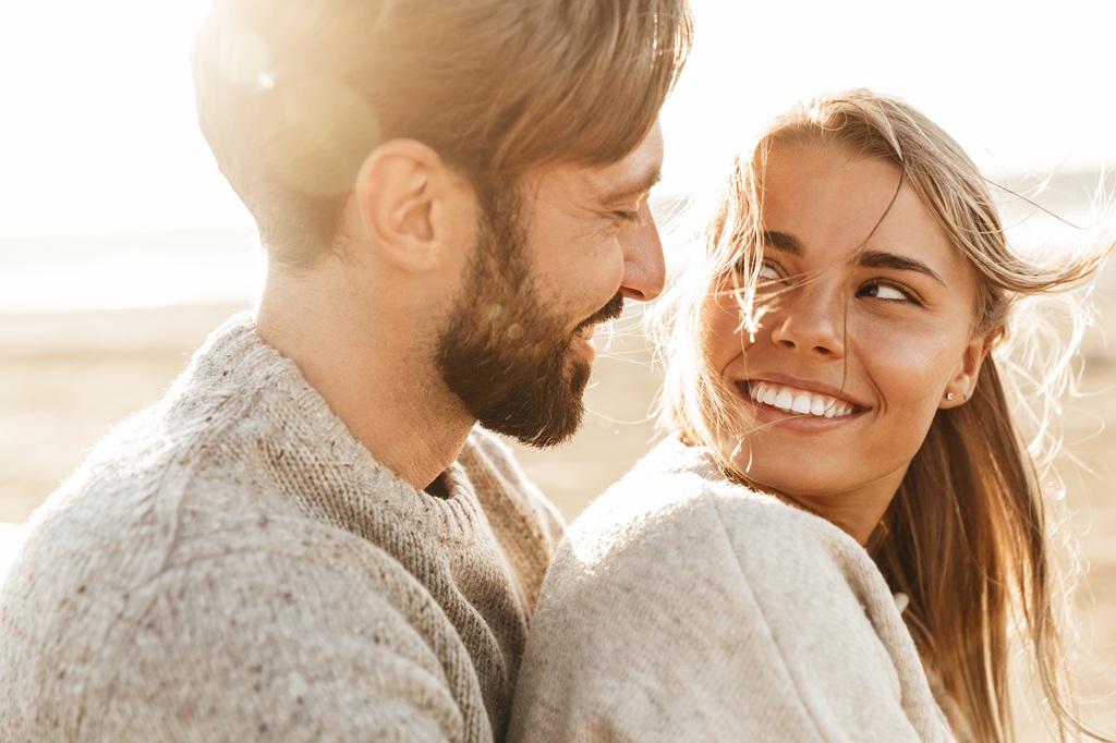 Close-up of man and woman in sweaters hugging from behind and looking into each other's eyes on the beach